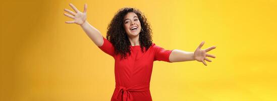 Come into my arms. Portrait of friendly and loving, caring charming woman with curly hair in red casual dress spread hands as wanting give hug smiling broadly at camera giving warm welcome or cuddle photo