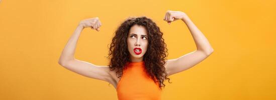 Woman feeling powerful and strong raising hands with clenched fists making intense face being working out in gym showing muscles and biceps looking at upper right corner posing over orange background photo