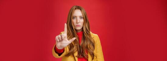 Serious-looking redhead woman in yellow coat extending hand to camera with stop and warning gesture prohibiting and forbidding make something having unemotional face as being confident over red wall photo