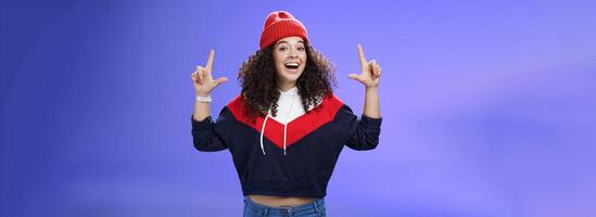 Waist-up shot of cheerful energized young woman calling friend inviting hang out as weather awesome pointing up with raised hands smiling broadly posing in red winter hat over blue background photo