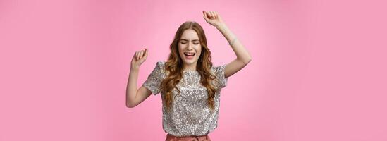 Attractive glamour female dancing dance-floor having fun rase hands up sky close eyes singing along songs enjoying awesome party cool music, wearing glittering blouse trendy skirt, pink background photo