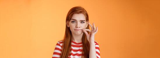 Girl can keep secrets. Serious-looking redhead female friend make promise not tell, seal mouth, zip lips gesture, look assertive and determined, stay silent, standing orange background photo