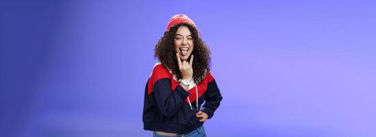 Stylish and cool energized swag girl with curly hairstyle in warm beanie showing rock-n-roll gesture near mouth and sticking out tongue smiling having fun, partying and enjoying life over blue wall photo