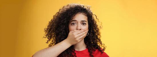 Close-up shot of concerned and insecure troubled woman keep silent scare of telling anyone her problem covering mouth with palm not to scream or slip word looking sad and sorrow over yellow background photo