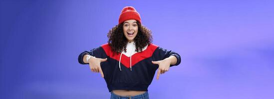Hurry up and chek it out. Portrait of charismatic joyful and excited young cute curly-haired girl in beanie and stylish outfit pointing down, smiling broadly inviting join fun over blue background photo