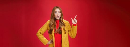 Shocked and stunned cute, confused ginger girl in yellow coat feeling worried and surprised open mouth lifting eyebrows amazed pointing at upper left corner, posing against red background photo