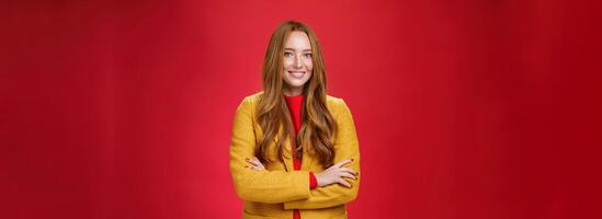 Charming successful young female in yellow autumn coat smiling broadly holding hands crossed on body in confident self-assured pose standing delighted and happy over red background photo