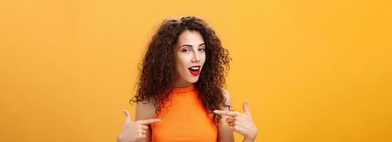 Proud and satisfied cool urban female with red lipstick and curly hairstyle pointing at herself with self-assured expression winking bragging about skills and achievements over orange background photo