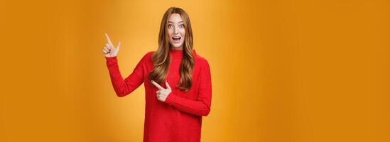 Enthusiastic and ambitious cute redhead female with freckles in red dress pointing at upper right corner smiling amazed and curious as exploring awesome new space posing over orange background photo