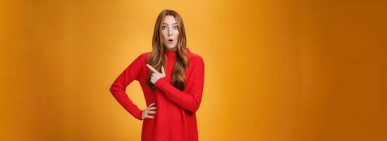 Impressed and surprised european ginger girl in red warm sweater open mouth from excitement and amazement pointing at upper right corner, gasping astonished and curious, asking question photo