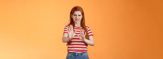 Gosh it stinks. Disgusted redhead picky woman blocking sign raise hands up defensive, grimacing, cringe from aversion awful smell, show refusal rejecting disgusting offer, stand orange background photo