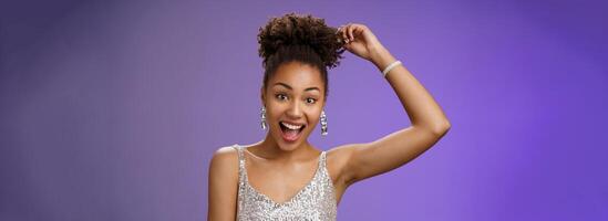 Close-up excited good-looking african-american curly-haired woman in trendy silver glittering dress touching afro curly smiling gladly having fun dancing amused standing entertained blue background photo