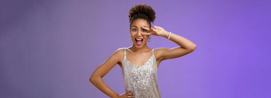 Charming lively african-american woman in glittering silver dress having fun smiling amused joyfully show victory peace gesture eye hold hand waist confident enthusiastic pose blue background photo