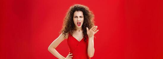 Girl chowing her temper to employees being fed up with unappropriate behaviour complaining standing pissed and dissatisfied gesturing with palm and holding hand on hip over red background photo