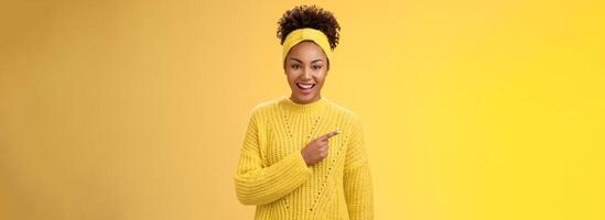 Friendly good-looking enthusiastic black charming girl in headband sweater smiling broadly amused pointing left awesome place showing you perfect blank space advertisement, posing yellow background photo