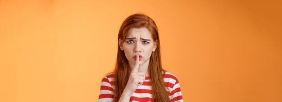 Uneasy worried insecure redhead girl begging keep secret safe, frowning grimace upset, shushing, press index finger lips hush gesture, worry someone spread rumor, orange background photo
