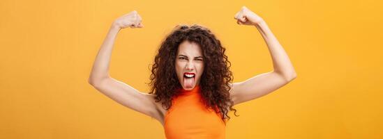 Waist-up shot of cool and daring caucasian female in orange top with tattoo on arm frowning making funny face sticking out tongue raising hands showing muscles feeling power and strengths photo