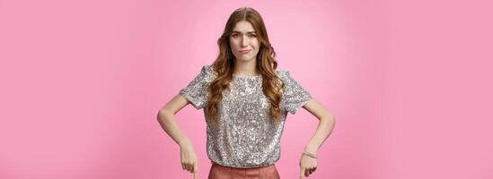 Well unimpressive. Girl gringe smirking frowning disappointment pointing down expressing displeased dislike, show bad uninteresting worthless product, being unamazed, standing pink background photo