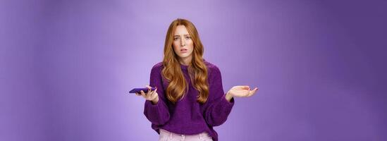 Clueless and sad cute redhead woman spread hands sideways frowning disturbed and upset holding smartphone being questioned and clueless why mobile phone not working, posing over purple wall photo
