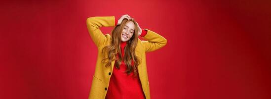Carefree happy ginger girl carrying away with music and good vibes holding hands on head close eyes and smiling delighted and relaxed having fun, dancing against red background in yellow coat photo