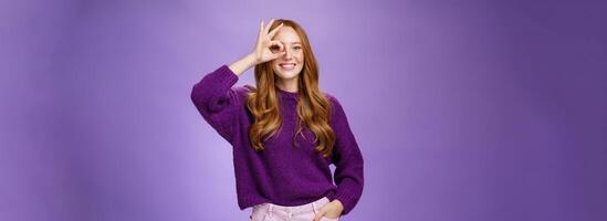 Optimistic girl looking into middle distance through okay gesture and smiling broadly with hopeful excited grin standing delighted over purple background in sweater and hand in pants photo