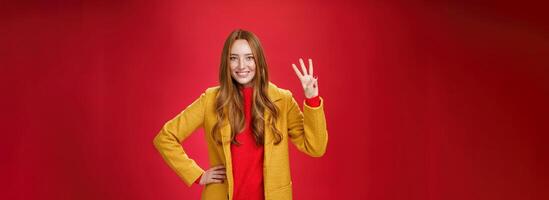 Cute friendly-looking ginger girl showing three reasons buy product smiling broadly making third reservation, looking delighted and confident in her choice posing against red background in yellow coat photo