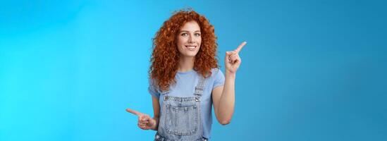 Lively joyful good-looking 25s redhead curly-haired sassy girl summer positive mood dancing overalls pointing sideways up right index fingers directing promos store links smiling delighted photo