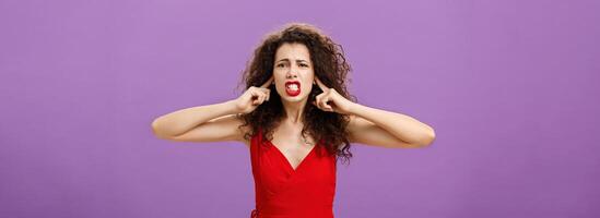 Woman fed up of hearing cursing words and arguments in her turn. Portrait of displeased insecure and offended female with curly hairstyle in red dress closing eyes and talking displeased and bothered photo