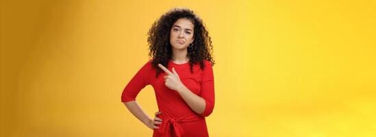 Portrait of unimpressed careless and bored young curly-haired female coworker in dress making upset smile pointing at upper right corner looking with indifference and disappointment photo