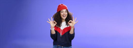 Got under control. Portrait of happy charming smiling curly-haired female in warm winter hat and sweatshirt smiling broadly and showing okay or excellent gesture as approving, liking cool movements photo