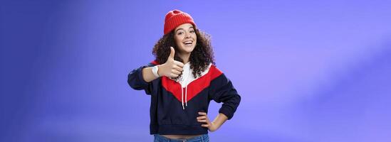Nice work, keep up. Delighted and supportive charming sportswoman in winter beanie, sweatshirt and watch showing thumbs up with satisfied smile, giving approval, liking and cheering great effort photo