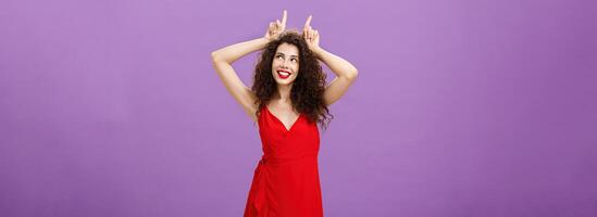 Woman being daredevil on party standing sensual and seductive in red elegant evening dress holding index fingers on head like devil horns looking at upper right corner with flirty smile photo
