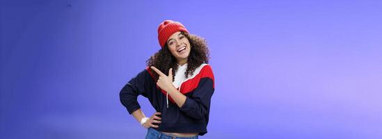Studio shot of enthusiastic charming curly-haired woman inviting to play in snow with her pointing at upper right corner tilting head joyfully and smiling, holding hand on waist over blue wall photo