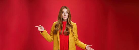 Confused and questioned cute redhead girl in yellow autumn coat pointing right as shrugging making clueless gesture with hand open mouth wondered as asking question over red background photo