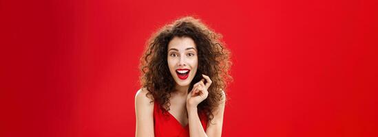 Impressed and excited feminine adult woman with curly hair in red lipstic and elegant evening dress gasping and smiling with surprised, amazed look playing with hair strand listening carefully story photo