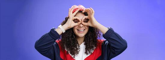Girl see us as imitating peek with binocular making circles over eyes and looking through it joyful and excited, having fun fooling around posing happy and cute over blue background in outdoor outfit photo