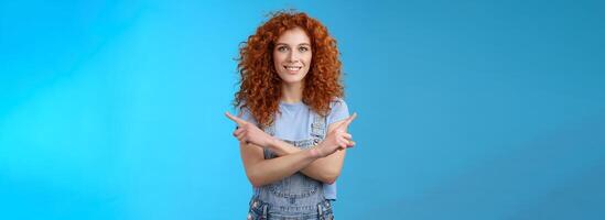 Which way. Charismatic attractive happy smiling redhead curly girl pointing curious sideways cross arms body indicating left right intrigued what choose making decision standing blue background photo