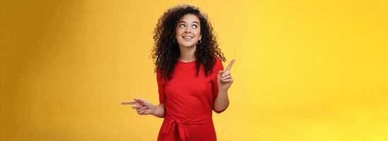 Dreamy charming woman making choice as checking out two awesome products pointing sideways looking intrigued and curious at upper left corner, smiling dreamy and delighted over yellow wall photo