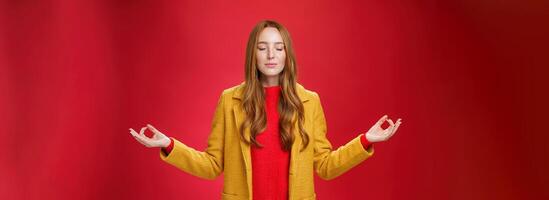 Girl keep calm releasing stress with meditation, posing in yellow coat, close eyes and looking relieved as extending hands sideways with mudra gesture, doing yoga against red background in lotus pose photo