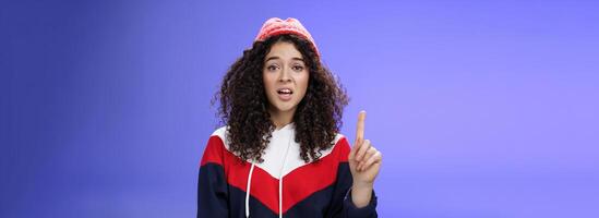 Rule number one do not mess with me babe. Portrait of swag and cool stylish young curly-haired woman in winter beanie showing index finger in prohibition or warning gesture over blue background photo