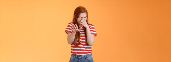 Gosh it stinks. Reluctant disgusted redhead woman raise hand block dislike gesture shut nose, cringe from aversion dislike, puke from reek bad smell, stand orange background disappointed photo