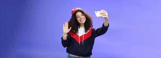 Studio shto of stylish cute female vlogger with curly hair in winter red hat smiling and waving hello at smartphone camera as recording , making interesting content to post online over blue wall photo