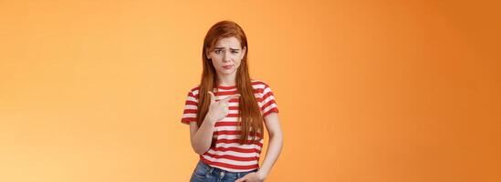 Upset uneasy cute ginger gitl long natural red hair, frowning smirking hesitant, complain, looking suspicious and doubtful point left, not sure if product really good, feel unsure, orange background photo
