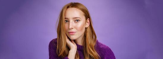 Sensual and romantic woman with red hair and freckles touching cheek gently as curls dropping on half face gazing charmed and flirty at camera, smiling being tender and coquettish over purple wall photo