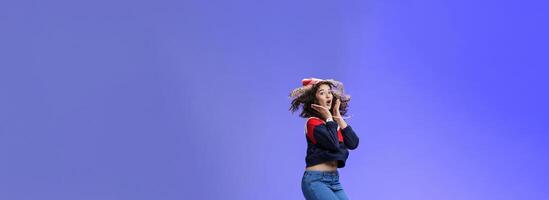 Surprised and amazed amused good-looking woman with curly hair in winter beanie and sweatshirt open mouth from joy and amazement jumping over blue background having fun feeling playful photo