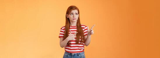 Complicated troubled cute young redhead girl puzzled facing tough difficult decision, look uneasy frowning perplexed hesitating, stare pointing upper left corner disappointed, orange background photo