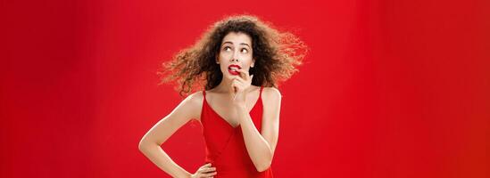 Waist-up shot of concerned and worried insecure cute female with curly hairstyle in red evening dress biting finger looking troubled or questioned at upper right corner with hand on waist photo