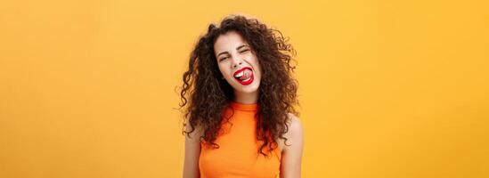 Portrait of upbeat playful and stylish caucasian girl with curly hair and red lipstick winking showing tongue with rebellious hot expression tilting head standing cool and chill over orange background photo