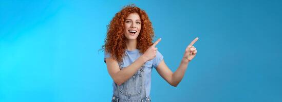 Hey check this out. Enthusiastic redhead curly stylish summer girl inviting beach party pointing upper left corner directing awesome location smiling broadly discuss awesome promo blue background photo