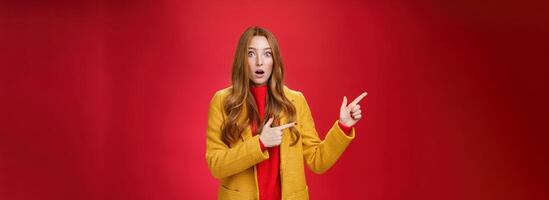 Stunned speechless and impressed attractive redhead woman with freckles in yellow coat drop jaw amazed as popping eyes at camera questioned, pointing left astonished over red background photo